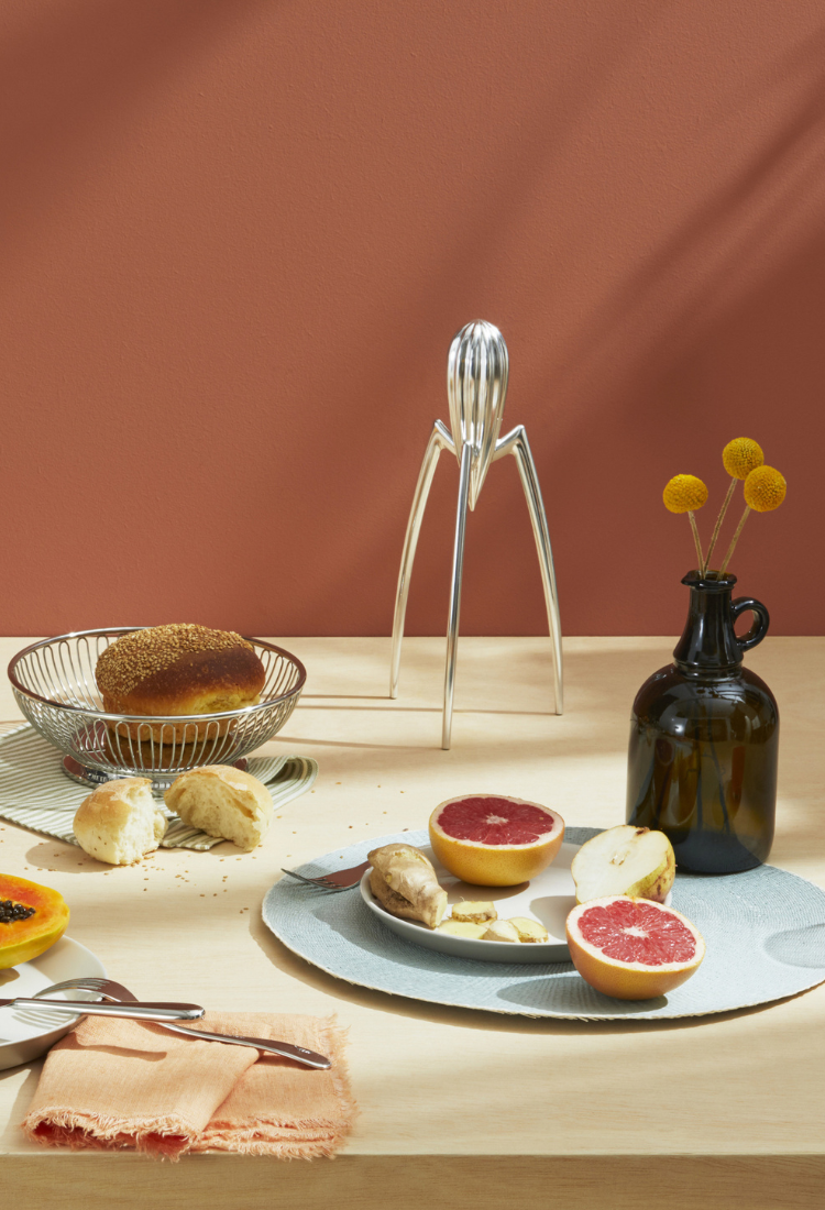 Gedeckter Frühstückstisch mit Produkten des Herstellers Alessi, links ein Pulcina Kaffeekocher dahinter ein Obstkorb mit Orangen sowie weißes Geschirr und einer Zitruspresse von Alessi