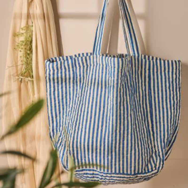 Gestreifte Stepp-Tasche in Off-White und Blau hängt an einem Haken vor beigem Hintergrund, dekorativ inszeniert mit Stoff und Pflanzen.