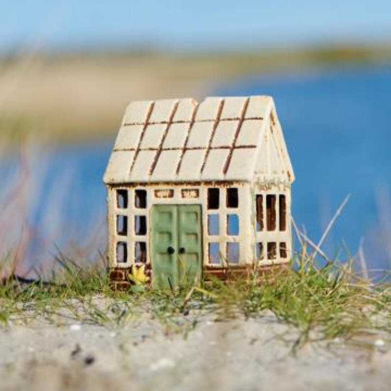 Deko-Teelichthaus mit grünem Tor und gelber Blume steht auf einer Sanddüne mit Blick auf ein Gewässer